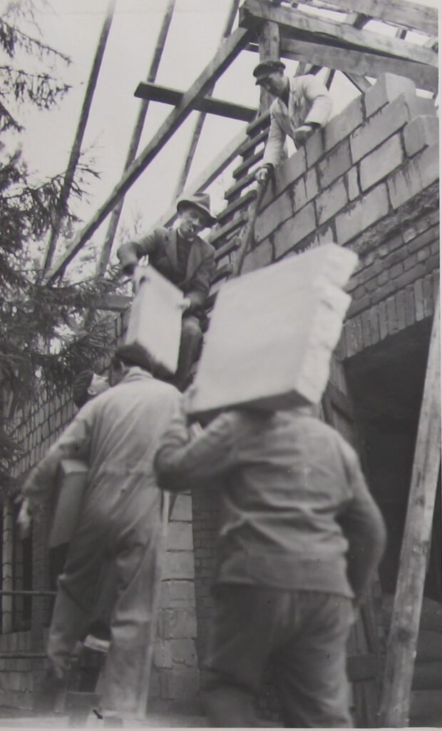 Foto schwarz-weiß aus den 50er Jahren, 2 Männer tragen Ziegelsteine vor einem Rohbau